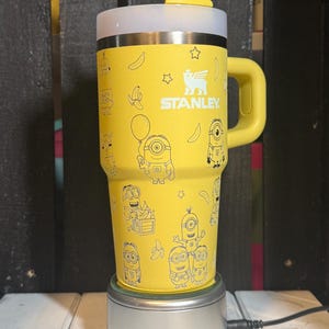 May include: A bright yellow Stanley tumbler featuring a repeating pattern of Minion characters, bananas, and stars in a thin black line art style. The cup includes a matching yellow handle, a white lid, and a clear straw protruding from the top.