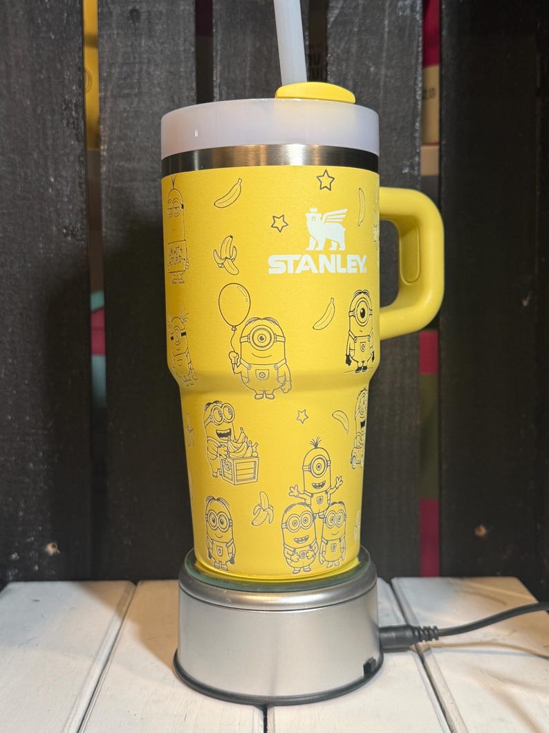 May include: A bright yellow Stanley tumbler featuring a repeating pattern of Minion characters, bananas, and stars in a thin black line art style. The cup includes a matching yellow handle, a white lid, and a clear straw protruding from the top.