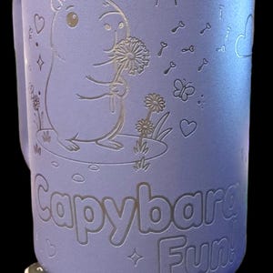 May include: A lilac travel mug featuring a capybara illustration. The design includes a sun, keys, butterflies, and hearts. The capybara holds a dandelion. The words "Capybara Fun!" are etched on the mug.