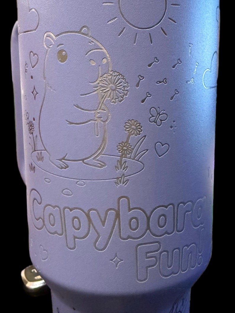 May include: A lilac travel mug featuring a capybara illustration. The design includes a sun, keys, butterflies, and hearts. The capybara holds a dandelion. The words "Capybara Fun!" are etched on the mug.