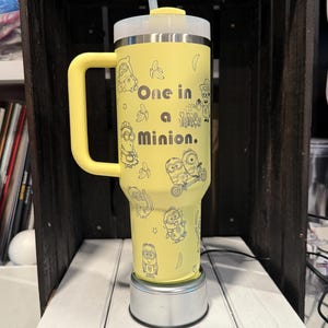 Pu&ograve; includere: Un bicchiere termico giallo con manico, coperchio trasparente e cannuccia. L'esterno presenta illustrazioni a tratto nero di personaggi Minion, banane e stelle, con il testo "One in a Minion." stampato al centro del bicchiere.
