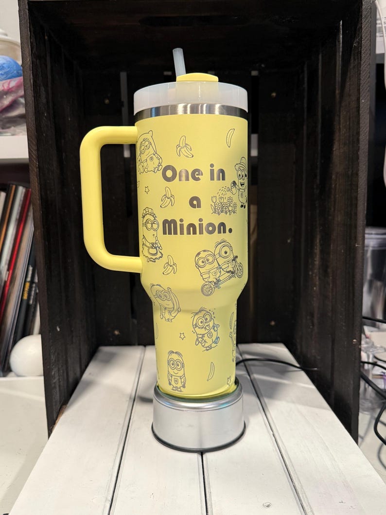 Pu&ograve; includere: Un bicchiere termico giallo con manico, coperchio trasparente e cannuccia. L'esterno presenta illustrazioni a tratto nero di personaggi Minion, banane e stelle, con il testo "One in a Minion." stampato al centro del bicchiere.