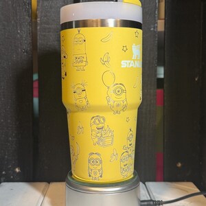 May include: A yellow Stanley tumbler featuring a repeating pattern of Minion characters, bananas, and stars in black line art. The cup has a white lid with a yellow straw cover and the text "IM WITH STUPID" printed on one of the Minion figures.