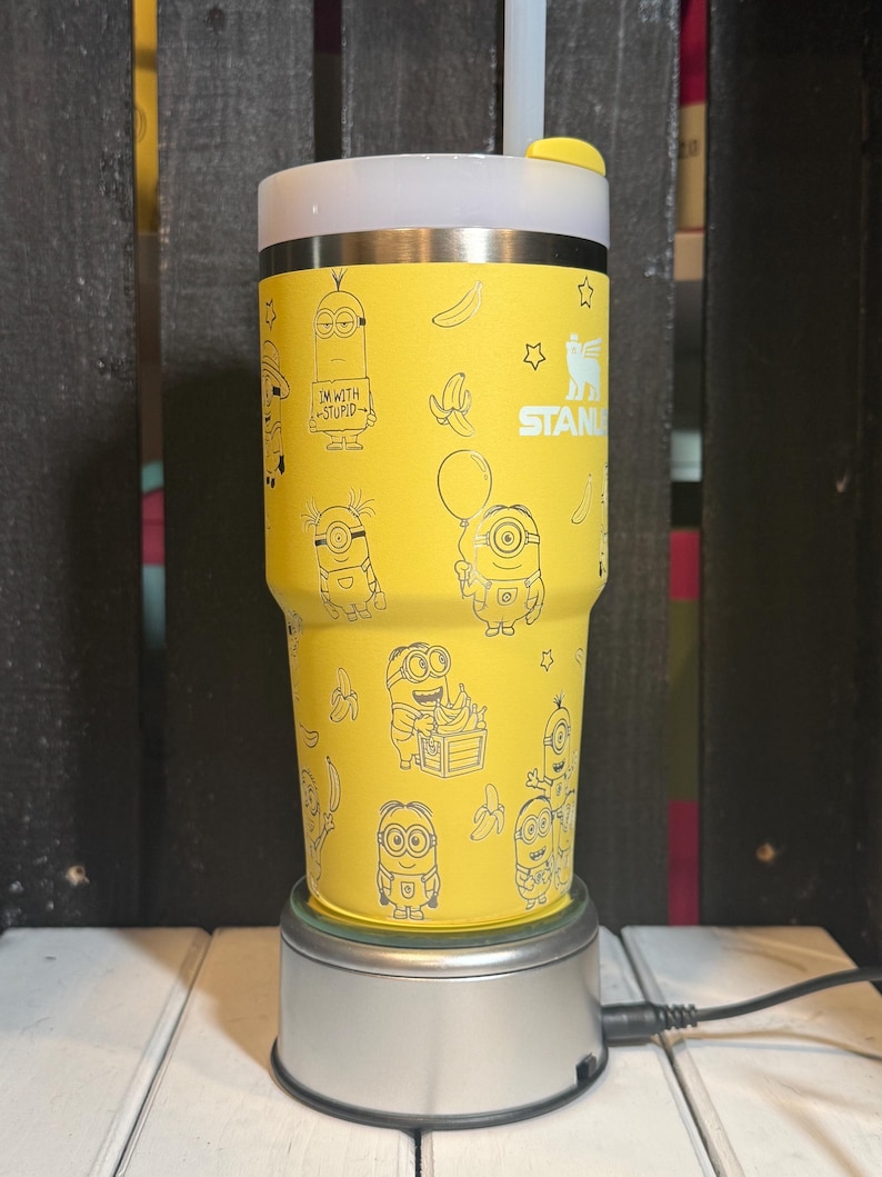 May include: A yellow Stanley tumbler featuring a repeating pattern of Minion characters, bananas, and stars in black line art. The cup has a white lid with a yellow straw cover and the text "IM WITH STUPID" printed on one of the Minion figures.
