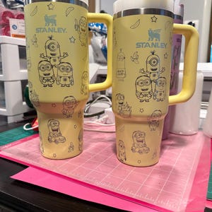 Pu&ograve; includere: Due bicchieri Stanley gialli con manici e coperchi bianchi, ciascuno caratterizzato da un motivo a linee nere di personaggi Minion, banane e stelle. I bicchieri hanno una cannuccia inserita nella parte superiore e il logo Stanley sul davanti.