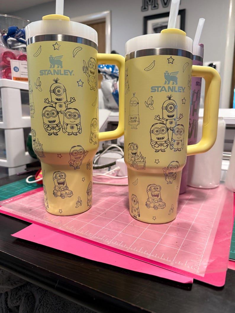Pu&ograve; includere: Due bicchieri Stanley gialli con manici e coperchi bianchi, ciascuno caratterizzato da un motivo a linee nere di personaggi Minion, banane e stelle. I bicchieri hanno una cannuccia inserita nella parte superiore e il logo Stanley sul davanti.