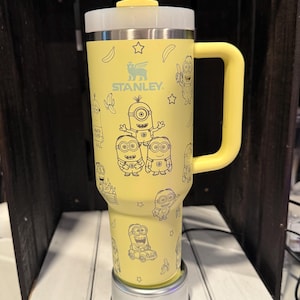 Pu&ograve; includere: Un bicchiere termico giallo Stanley con manico, cannuccia trasparente e coperchio. L'esterno presenta un motivo ripetuto a linee nere con personaggi Minion, banane e stelle. Il logo Stanley &egrave; stampato in nero nella parte superiore del bicchiere.