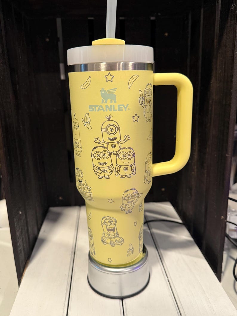 Pu&ograve; includere: Un bicchiere termico giallo Stanley con manico, cannuccia trasparente e coperchio. L'esterno presenta un motivo ripetuto a linee nere con personaggi Minion, banane e stelle. Il logo Stanley &egrave; stampato in nero nella parte superiore del bicchiere.