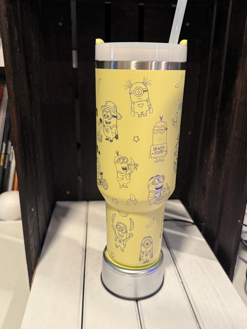 Pu&ograve; includere: Un bicchiere giallo con coperchio bianco e cannuccia trasparente, caratterizzato da un motivo ripetuto a linee nere di personaggi Minion e banane. Un Minion indossa un cappello, mentre un altro tiene un cartello con scritto I'M WITH STUPID.