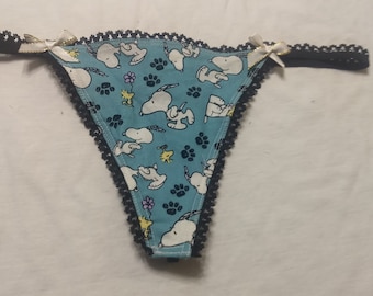 Puede incluir: Tanga azul claro con un estampado de Snoopy y Woodstock. Los bordes están adornados con encaje negro y pequeños lazos blancos con detalles dorados. La tela tiene un patrón repetido de Snoopy, huellas de patas y flores.