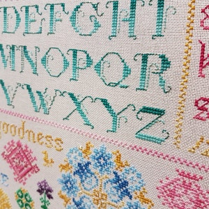 May include: Close-up of a cross-stitch sampler featuring a teal alphabet, the word "goodness" stitched in yellow, and colourful floral designs.