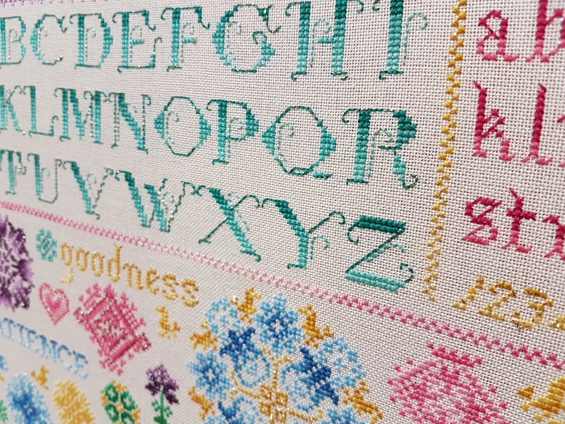 May include: Close-up of a cross-stitch sampler featuring a teal alphabet, the word "goodness" stitched in yellow, and colourful floral designs.