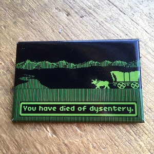 Puede incluir: Un imán negro y verde con una imagen pixelada de una carreta cubierta y una vaca. El texto "You have died of dysentery." se muestra en verde.
