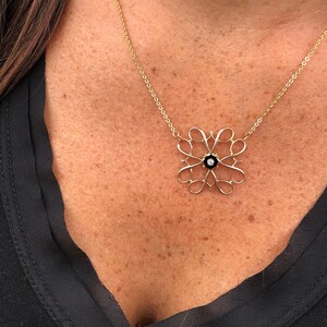 Pu&ograve; includere: Una collana con catena d'oro con un pendente delicato e intricato con un centro in smalto nero e un piccolo diamante. Il pendente ha la forma di un fiore stilizzato con pi&ugrave; anelli.