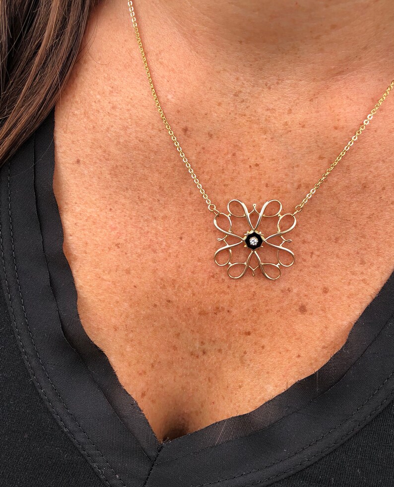 Pu&ograve; includere: Una collana con catena d'oro con un pendente delicato e intricato con un centro in smalto nero e un piccolo diamante. Il pendente ha la forma di un fiore stilizzato con pi&ugrave; anelli.