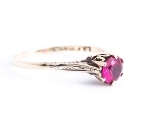 May include: A gold ring with a single, round, red gemstone set in a decorative, ornate setting.