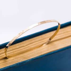 May include: A gold hammered metal bangle bracelet, lying on a blue and yellow book.