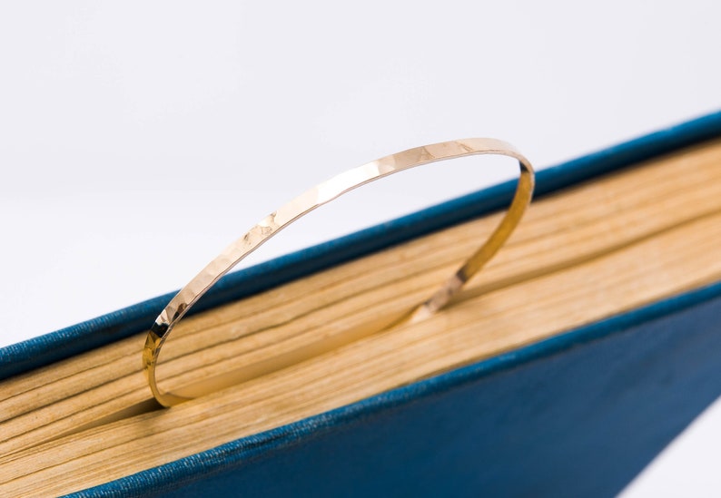 May include: A gold hammered metal bangle bracelet, lying on a blue and yellow book.