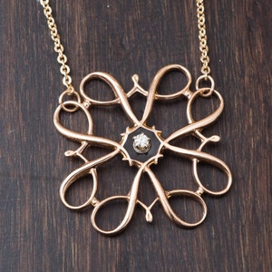Pu&ograve; includere: Una collana d'oro con un pendente delicato e intricato che presenta un centro in smalto nero con un solo diamante. Il pendente ha la forma di un fiore stilizzato con pi&ugrave; anelli e volute.