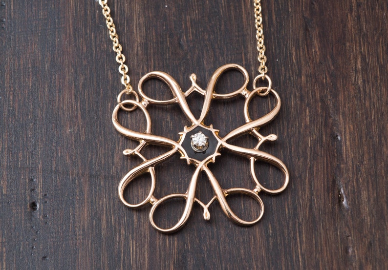 Pu&ograve; includere: Una collana d'oro con un pendente delicato e intricato che presenta un centro in smalto nero con un solo diamante. Il pendente ha la forma di un fiore stilizzato con pi&ugrave; anelli e volute.