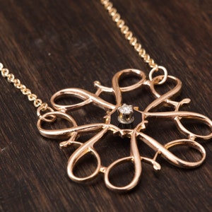 Pu&ograve; includere: Una collana d'oro con un pendente delicato e intricato con un singolo diamante incastonato in un centro nero. Il pendente ha la forma di un fiore stilizzato con pi&ugrave; anelli e curve.