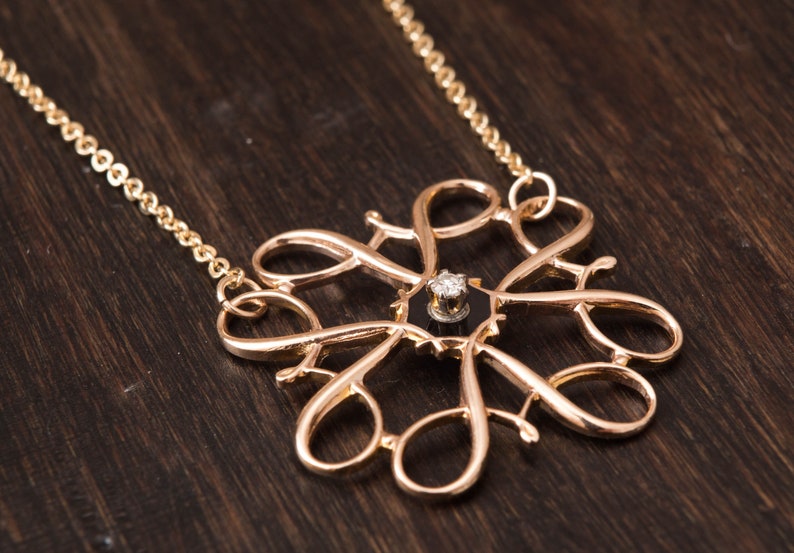 Pu&ograve; includere: Una collana d'oro con un pendente delicato e intricato con un singolo diamante incastonato in un centro nero. Il pendente ha la forma di un fiore stilizzato con pi&ugrave; anelli e curve.