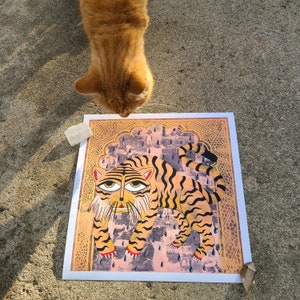 Peut inclure: Une illustration color&eacute;e d'un tigre avec un motif ray&eacute;, sur fond de ville rose et jaune. Le tigre est repr&eacute;sent&eacute; dans une pose ludique, la langue tir&eacute;e. L'illustration est encadr&eacute;e d'une bordure dor&eacute;e.