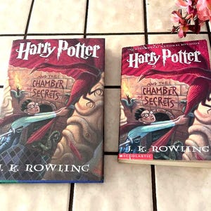 May include: Two copies of the book Harry Potter and the Chamber of Secrets by J.K. Rowling. The covers feature a red and brown illustration of a character with glasses reaching toward a large red bird in a stone chamber with green snakes.