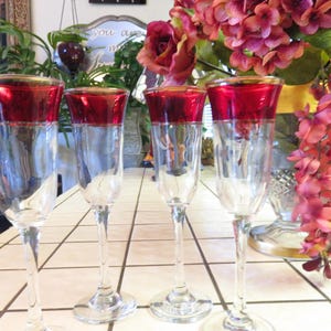 May include: Four clear champagne flutes with a red rim and gold trim. The flutes have a long stem and a wide base. The glasses are arranged on a white tiled surface. The background includes flowers and greenery.