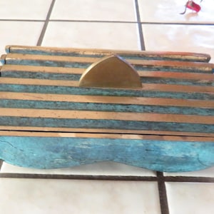 May include: A rectangular box with a lid, featuring a unique design. The box is made of metal with a combination of bronze and teal colors. The lid has a bronze, half-moon shaped handle.