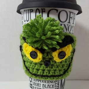 May include: A ceramic coffee mug with a black lid, featuring a green crocheted sleeve. The sleeve is designed to resemble a cartoon character's face, with yellow eyes, black button pupils, and a green pom-pom on top. The mug has text that says "Coffee".