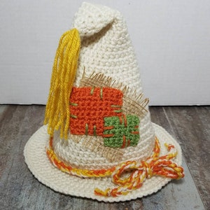May include: A white crocheted scarecrow hat with a yellow tassel, orange and green fabric patches, and a yellow and orange braided trim.
