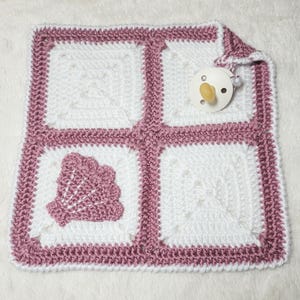 May include: A handmade crochet baby blanket in white and mauve. The blanket features a square design with a shell applique and a mauve border. A pacifier rests on the blanket. The blanket is made of soft yarn.