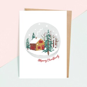 以下が含まれることがあります： 雪だるまの絵が描かれた白いクリスマスカード。雪だるまは、赤い窓と煙突のある小さな茶色の家と、緑の木々に囲まれています。雪だるまの下には、赤い筆記体で「メリークリスマス」と書かれています。