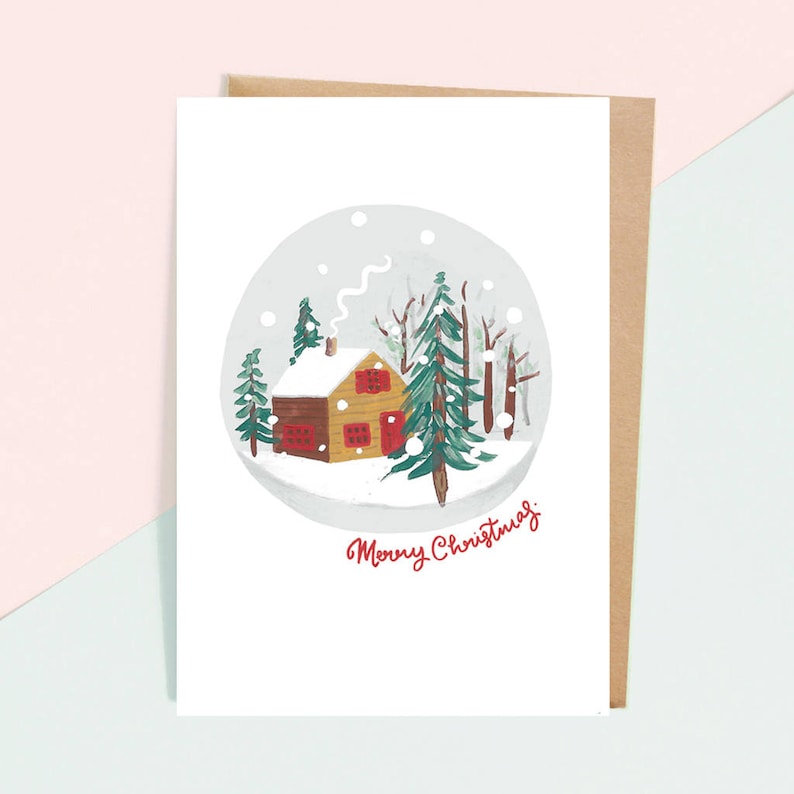 以下が含まれることがあります： 雪だるまの絵が描かれた白いクリスマスカード。雪だるまは、赤い窓と煙突のある小さな茶色の家と、緑の木々に囲まれています。雪だるまの下には、赤い筆記体で「メリークリスマス」と書かれています。