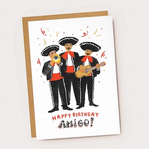 Puede incluir: Tarjeta de felicitación de cumpleaños con una ilustración en acuarela de tres músicos mariachis con trajes negros, detalles en rojo y sombreros grandes. La tarjeta dice "Happy Birthday Amigo!" con confeti. La tarjeta está sobre un respaldo de papel marrón.