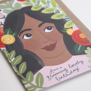 Puede incluir: Una tarjeta de cumplea&ntilde;os rosa y blanca con una ilustraci&oacute;n colorida de una mujer con flores en el pelo. El texto de la tarjeta dice "Have a Blooming Lovely Birthday!"