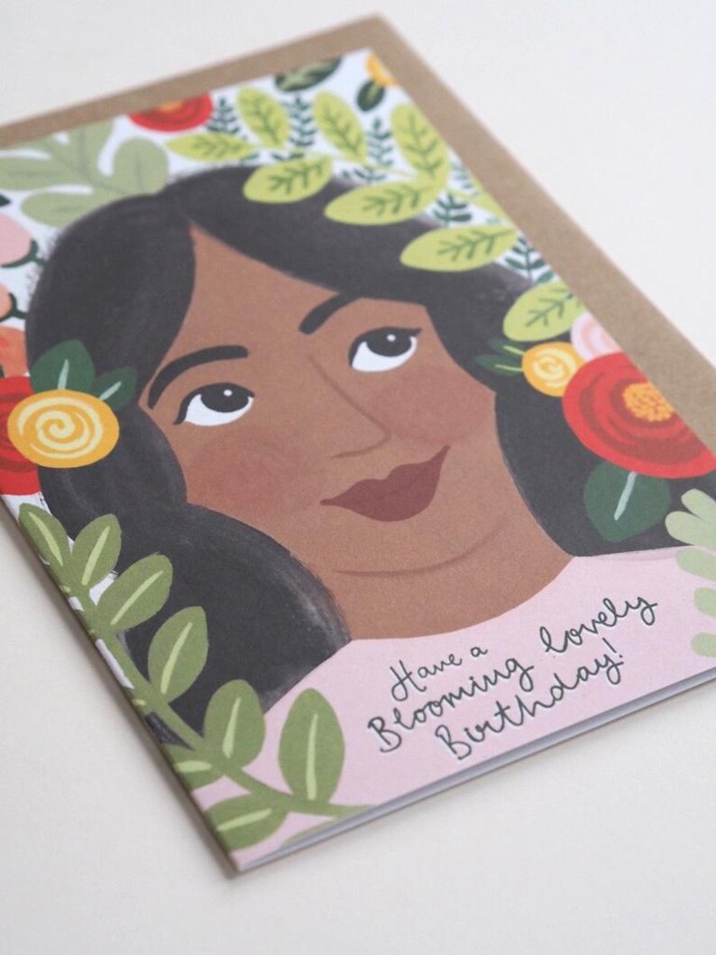 Puede incluir: Una tarjeta de cumplea&ntilde;os rosa y blanca con una ilustraci&oacute;n colorida de una mujer con flores en el pelo. El texto de la tarjeta dice "Have a Blooming Lovely Birthday!"