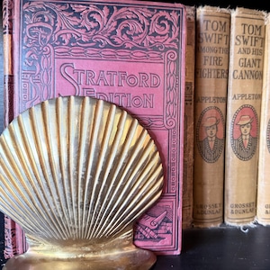 May include: A gold-toned metal seashell bookend positioned in front of a red Stratford Edition book, alongside a row of vintage Tom Swift books with tan spines and blue books with gold lettering on a shelf, creating a classic library display.