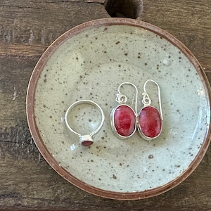 May include: A set of silver earrings with red oval gemstones and a silver ring with a red gemstone. The jewelry is displayed on a speckled gray ceramic dish.
