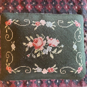 May include: A green needlepoint pillow with a floral design featuring pink, white, and blue flowers. The pillow has a white border with a decorative scroll pattern.