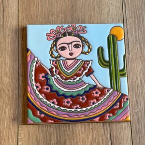 May include: A square ceramic tile featuring a colorful illustration of a person with braided hair and floral headwear, wearing a red dress with pink flower patterns. The background shows a light blue sky, a green cactus, and a yellow sun.