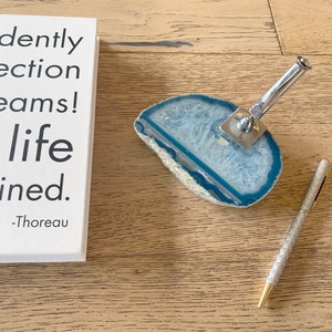 May include: A silver pen holder with a blue and white agate base. The pen holder is on a wooden surface. The text on the book reads: "Confidently in the direction of your dreams! The life you imagined. -Thoreau"