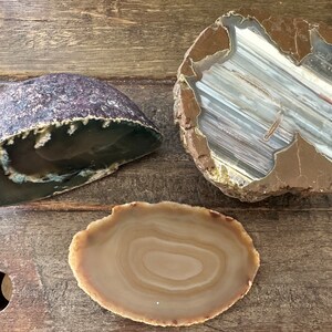 May include: Three agate slices with different patterns and colors. The first slice is a dark brown and purple agate with a rounded shape. The second slice is a light brown agate with a white and brown striped pattern. The third slice is a light brown agate with a circular pattern.