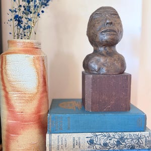 May include: A still life arrangement featuring a brown ceramic bust on a dark wooden block, stacked on vintage blue books. A tall, textured vase with dried blue flowers adds a touch of color and contrast to the scene.