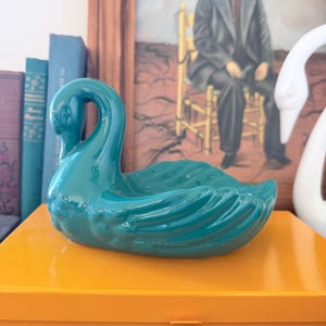 May include: A glossy teal ceramic swan figurine shaped as a shallow dish. The swan has a curved neck and detailed wing feathers molded into the body. The piece is finished with a smooth, reflective glaze and features a hollow center for storage.