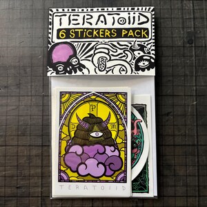 Può includere: Una confezione da 6 adesivi etichettata TERATOIID. L'intestazione presenta illustrazioni in bianco e nero di creature. L'adesivo frontale mostra una grafica viola e gialla di una figura cornuta con un occhio solo all'interno di una cornice ad arco, con il testo TERATOIID in basso.