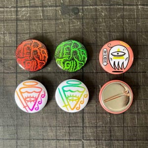 Puede incluir: Seis chapas redondas con alfiler que presentan varios diseños gráficos. Dos chapas tienen el texto "TERATOID" en rojo y verde con arte lineal negro. Tres chapas muestran un ojo estilizado con un halo. Una chapa muestra el mecanismo del alfiler.