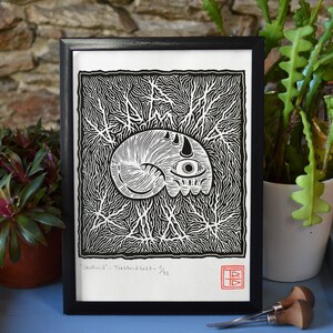 Peut inclure: Gravure sur linoléum en noir et blanc d'une créature avec un œil et une seule corne. L'impression est encadrée dans un cadre noir et porte le texte "Skulloid - Teratoid 2023 - 1/32" en bas à gauche.