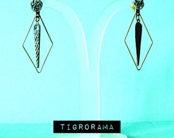 May include: A pair of geometric earrings on a clear display stand. Each earring features a diamond-shaped frame with a textured, silver-colored pendant. The earrings have a small, decorative top. The word "TIGORAMA" is displayed on a black rectangle.